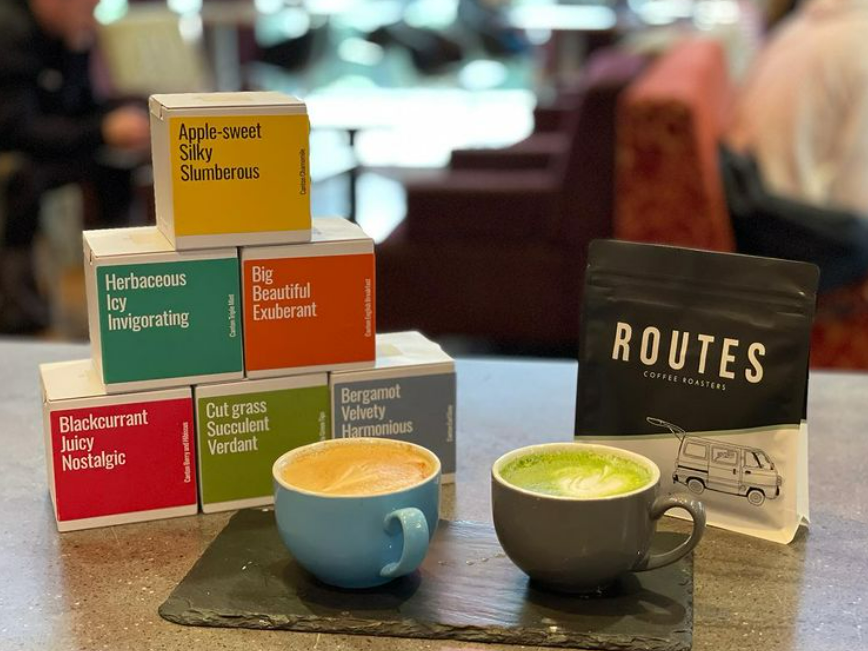 Small colourful boxes of Routes coffee are stacked next to two fresh cups of coffee